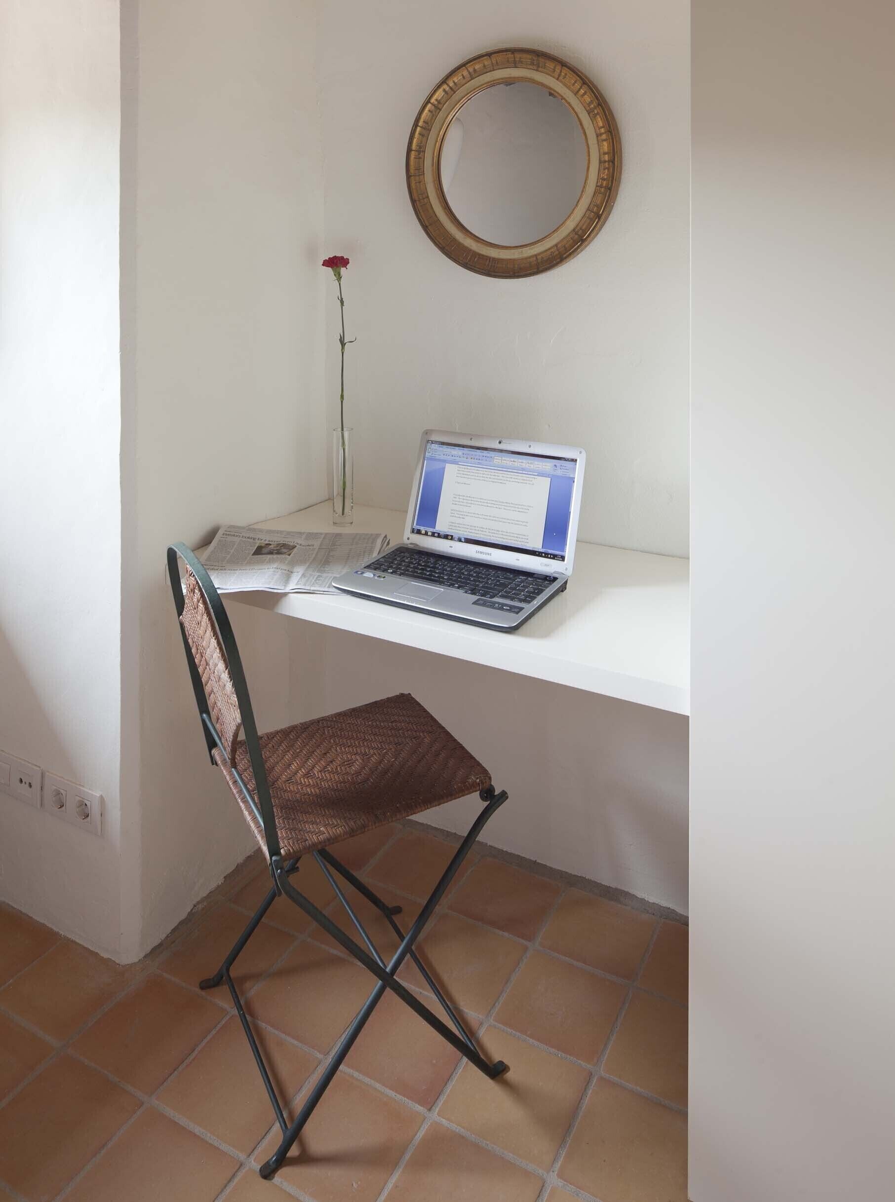Et lille arbejdsområde med en bærbar computer på et hvidt skrivebord, en fletstol, en vase med en enkelt blomst og et rundt spejl på væggen.