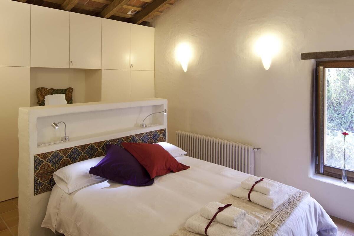 A bedroom with a bed, white walls, a wooden ceiling, a radiator, and a window with a view.