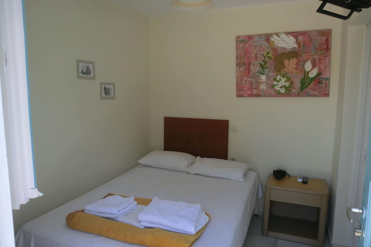 Una cama bien hecha con sábanas blancas y una toalla doblada, una mesita de noche con un control remoto y un cuadro en la pared.