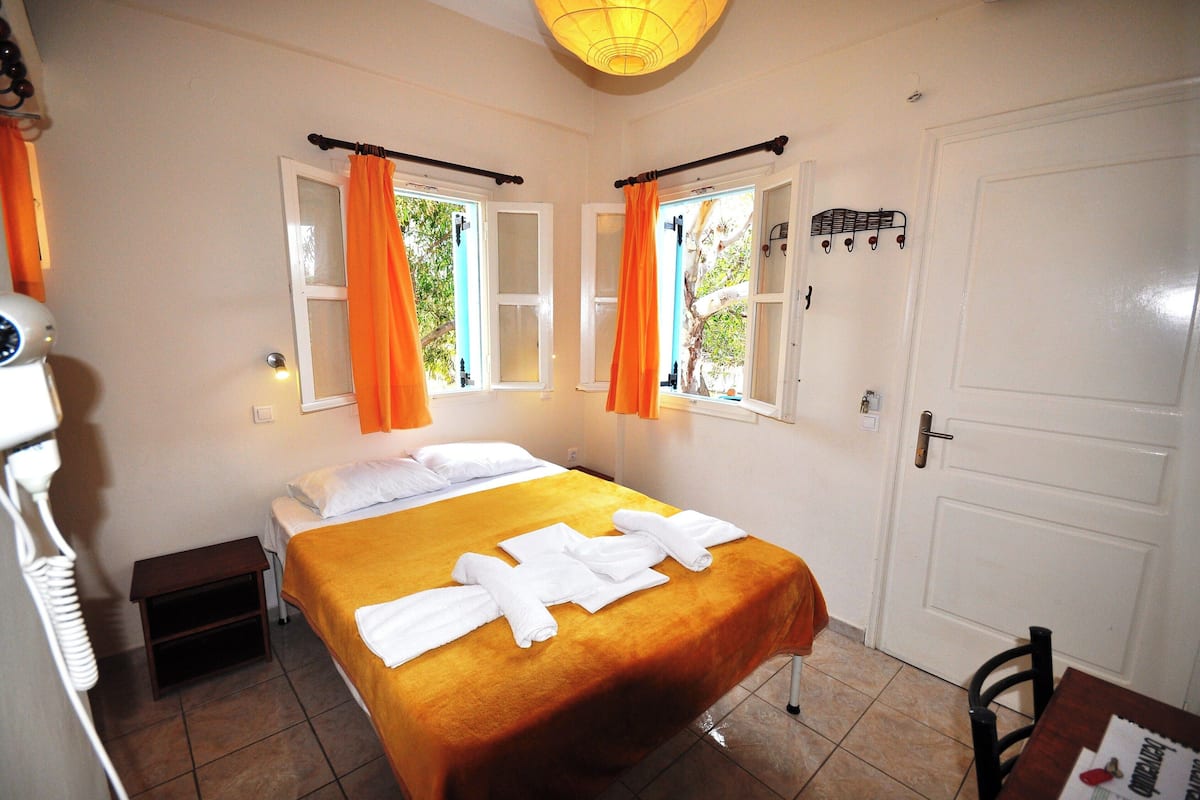 Habitación de hotel con cama, cortinas naranjas, una puerta blanca y una mesita con un periódico.