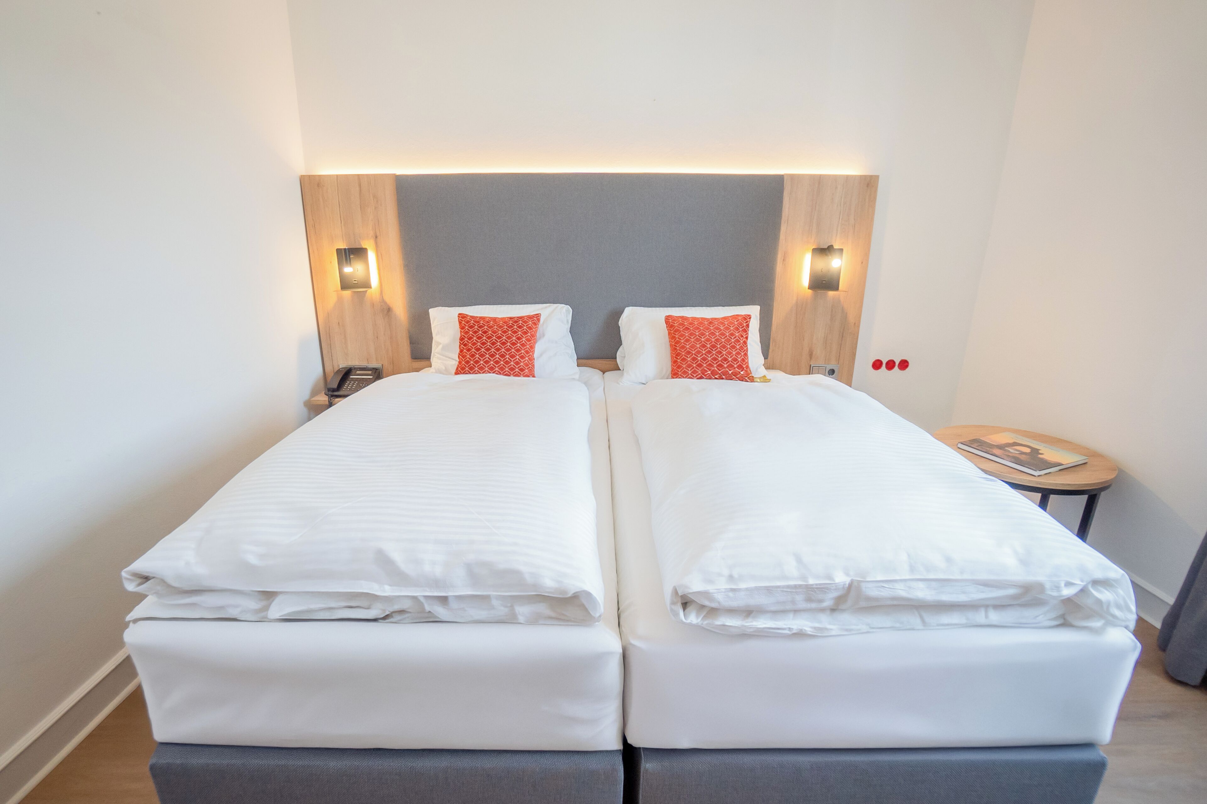 A double bed with white bedding and red pillows, a bedside table with a phone, and a small round table with a magazine.