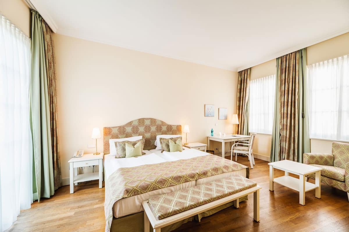 A hotel room with a bed, bedside tables, a desk, a chair, a bench, and curtains.