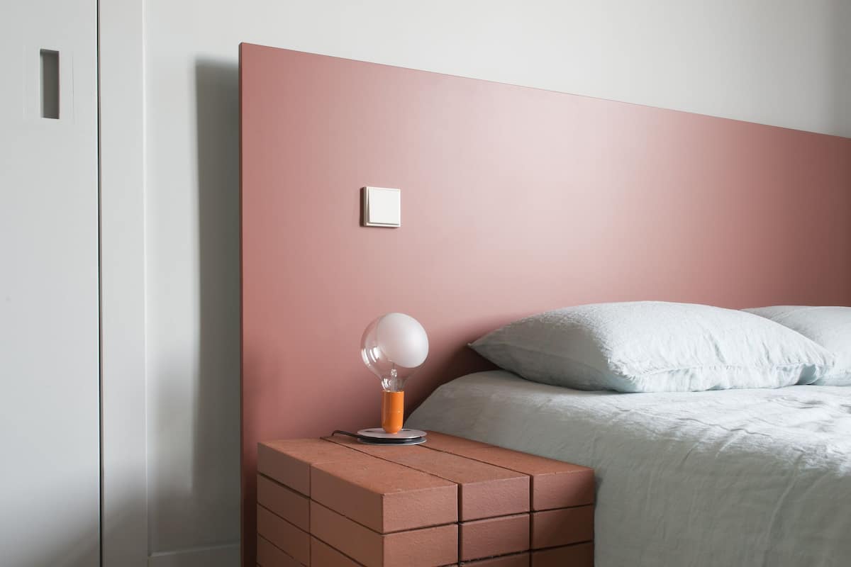 Un dormitorio con cabecera rosa, mesita de noche con lámpara y suelo de madera.