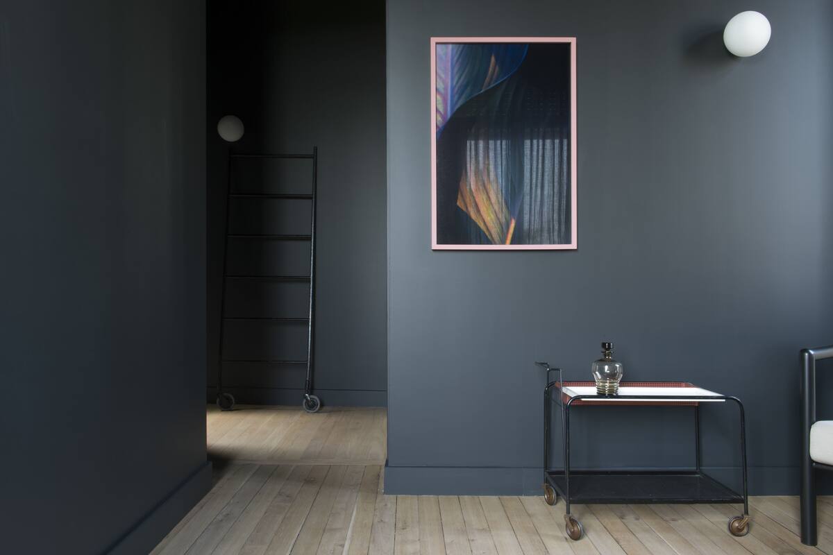 Una habitación con una pared gris oscuro, un cuadro abstracto enmarcado, una pequeña mesa con una botella, una silla y una escalera en la esquina.