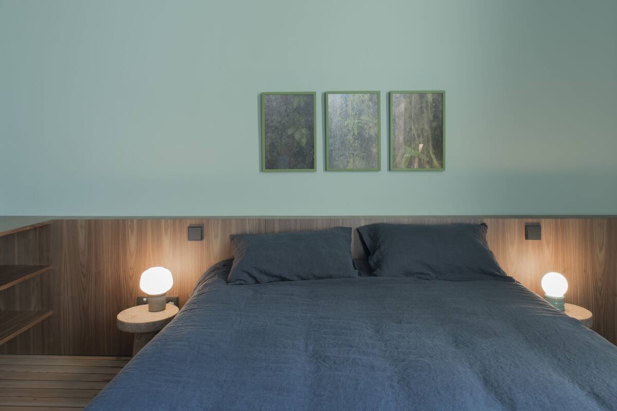 Un dormitorio con cabecera de madera, cama con ropa oscura, dos lámparas de noche y tres cuadros en la pared.