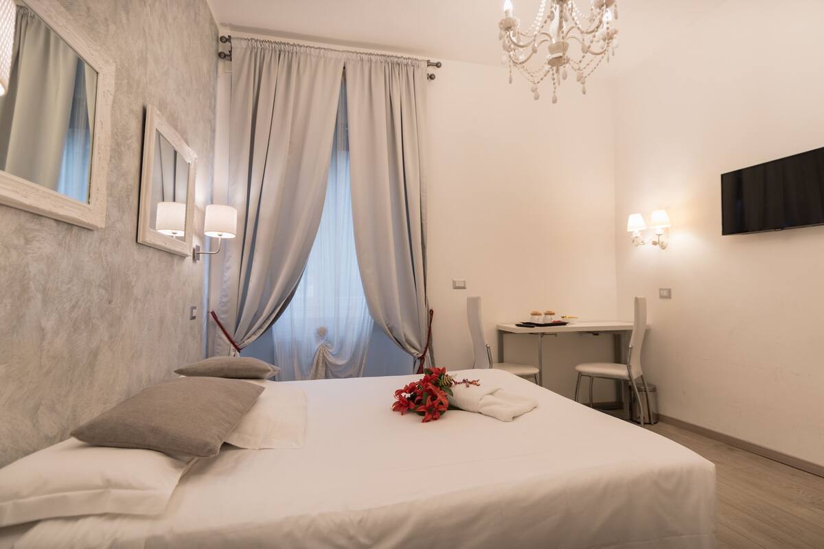 A hotel room with a bed, a desk, a chair, a TV mounted on the wall, and a chandelier.