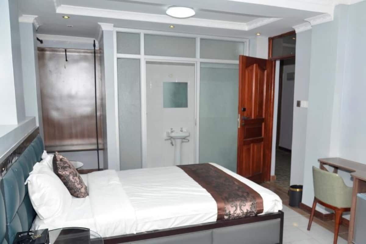 A hotel room with a bed, a desk, a chair, a TV, and a bathroom with a shower and toilet.