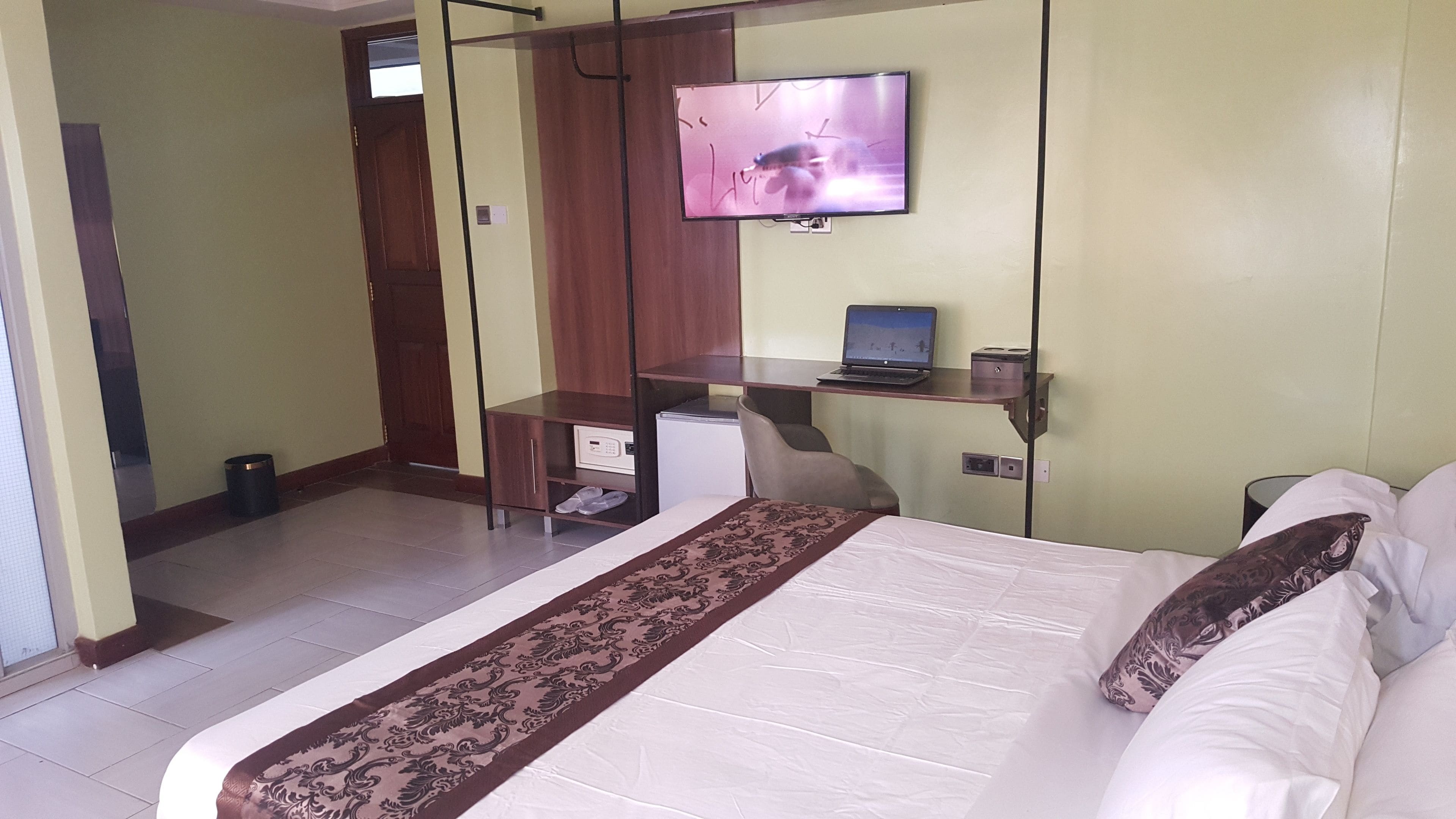 A hotel room with a bed, a desk with a laptop, a television, and a small refrigerator.