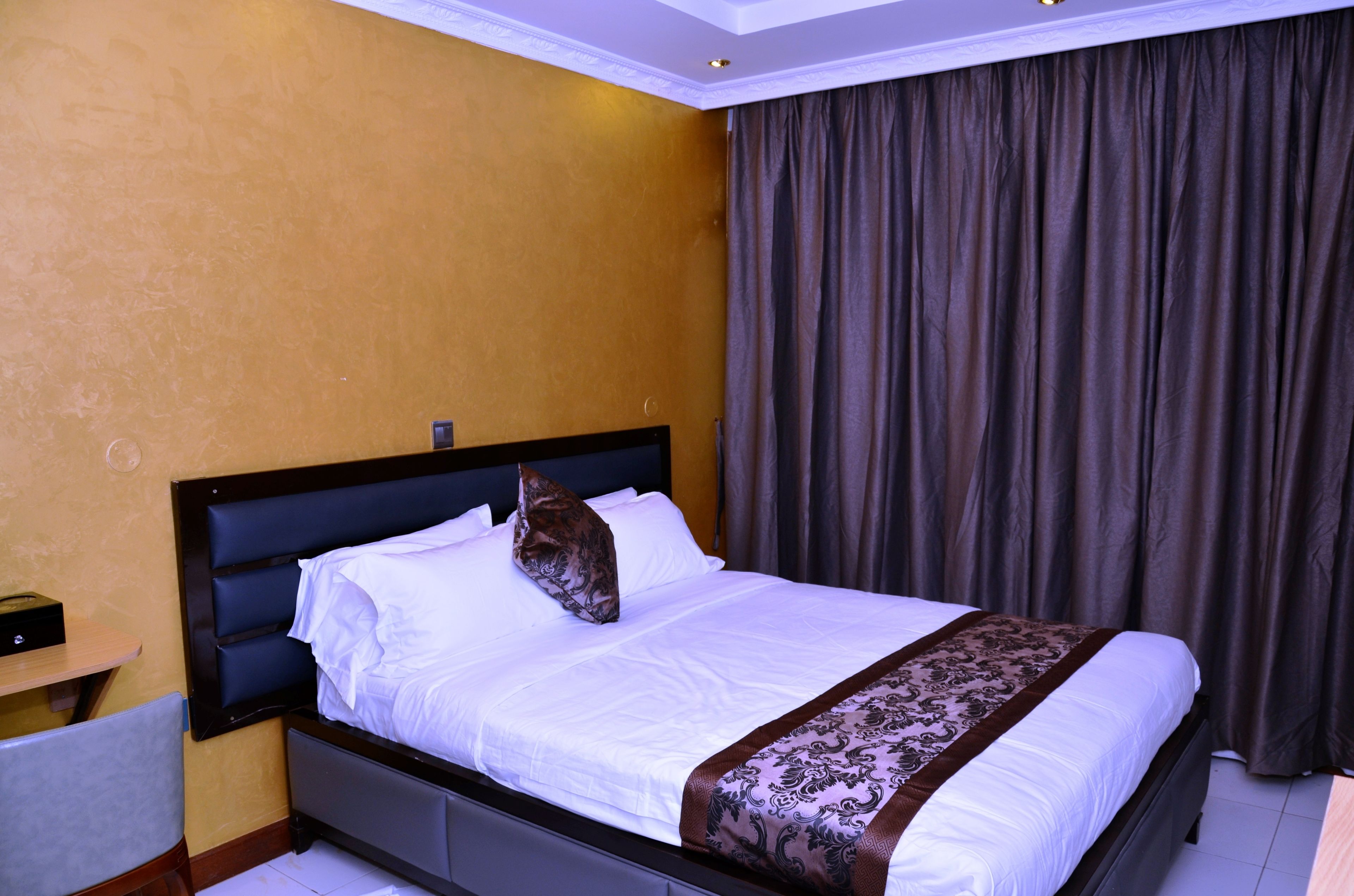 Una habitación de hotel con una cama, una mesita de noche, una silla y un amplio ventanal con cortinas.