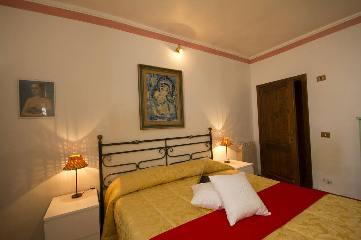 A neatly made bed with a red blanket, two bedside lamps, a framed painting, and a wooden door.