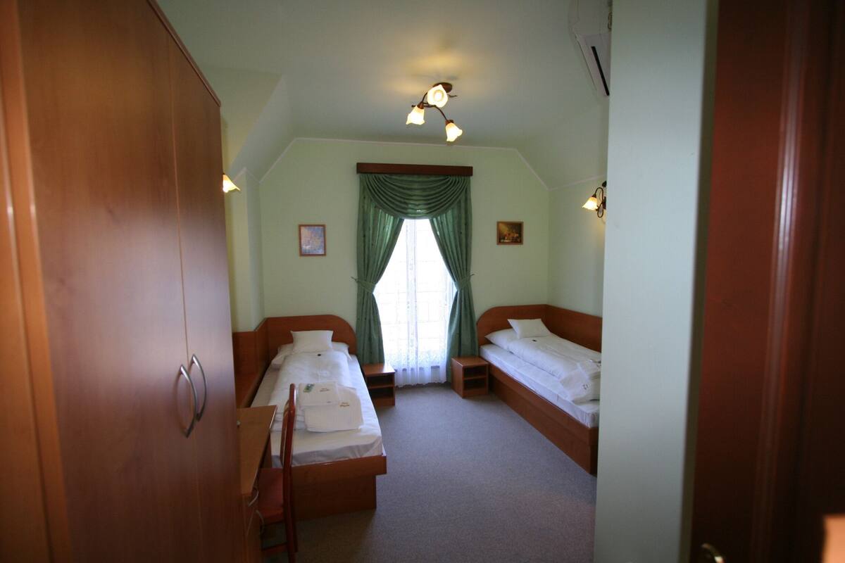 Ein Schlafzimmer mit zwei Einzelbetten, einem Holzschrank, einem kleinen Nachttisch und einem Fenster mit grünen Vorhängen.