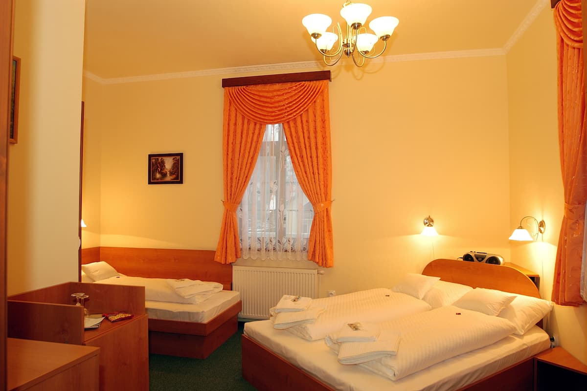 Ein Hotelzimmer mit Doppelbett, einem Holzkopfende, einem Nachttisch, einem Heizkörper und einer Deckenleuchte. Vor dem Fenster hängen orangefarbene Vorhänge.