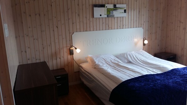 A bedroom with a wooden panel wall, a bed with white bedding, two bedside tables, and a wall-mounted light fixture.