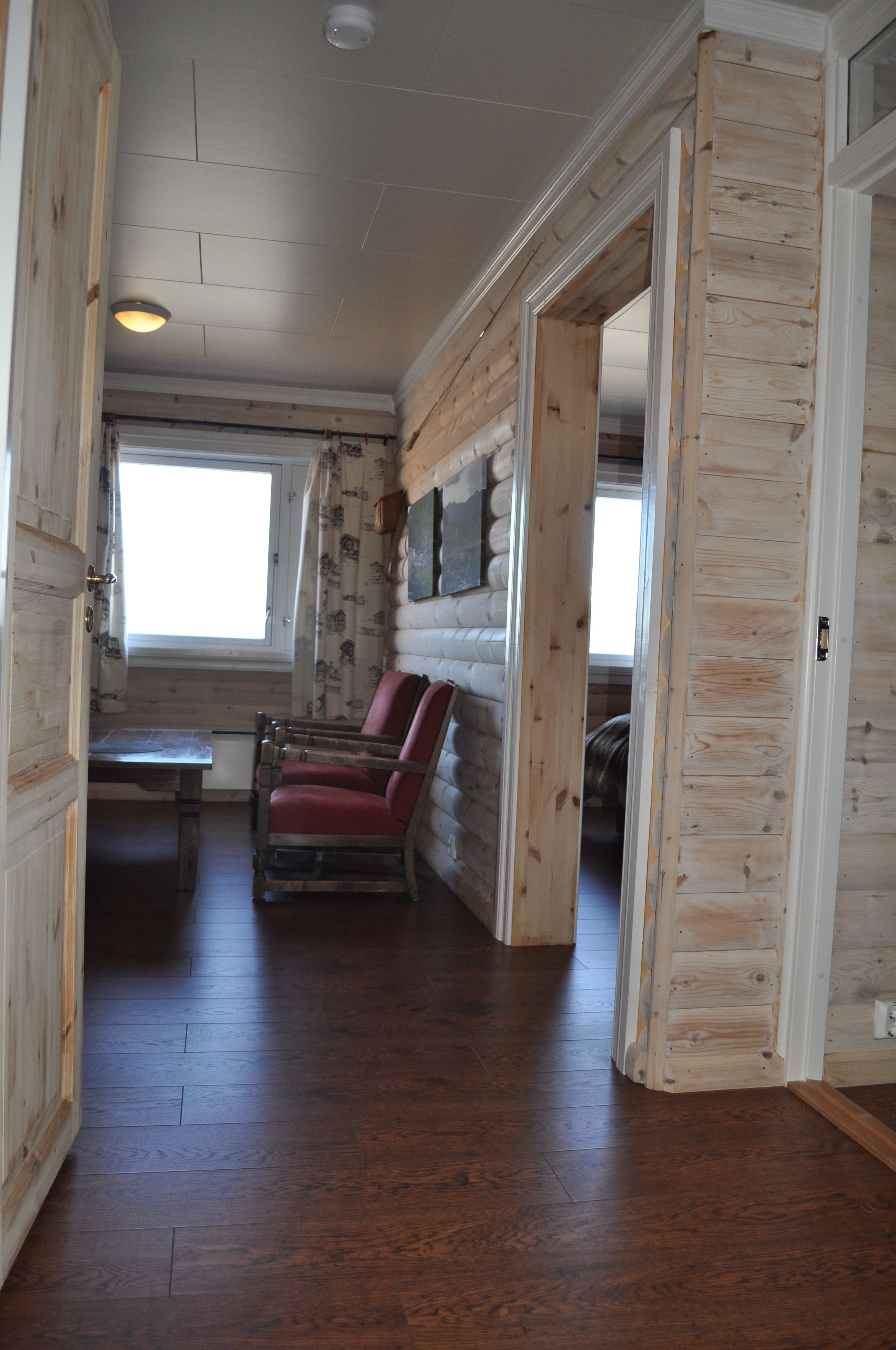 A log cabin interior with a wooden floor, a dining table, a rocking chair, and a window providing natural light.
