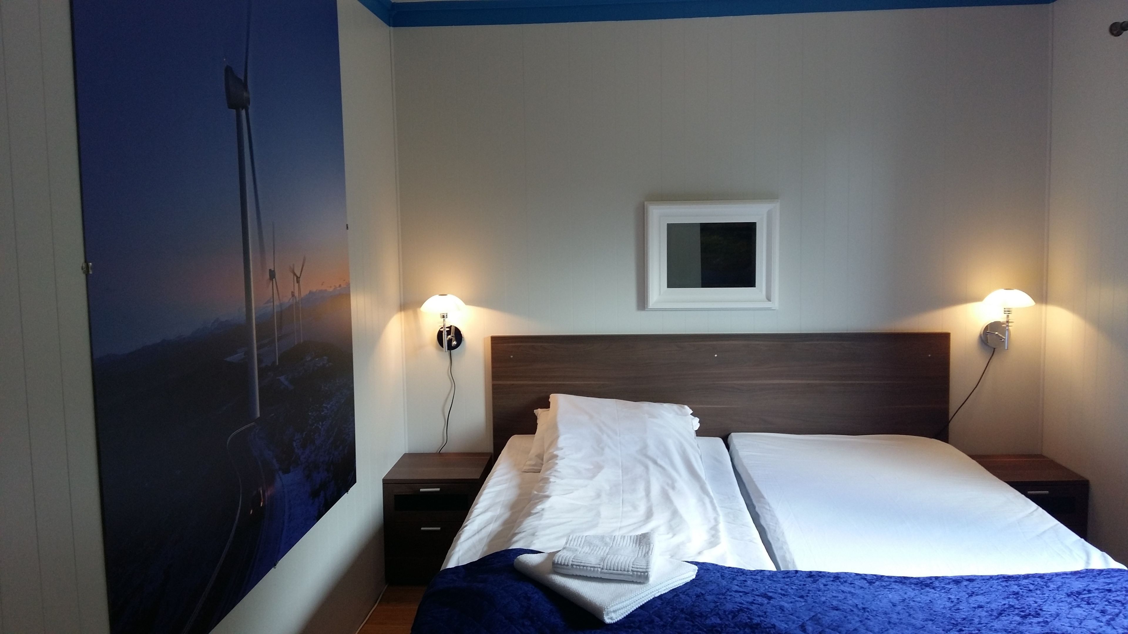 A hotel room with a bed, two bedside tables, a nightstand, a wall-mounted lamp, and a framed picture above the bed.