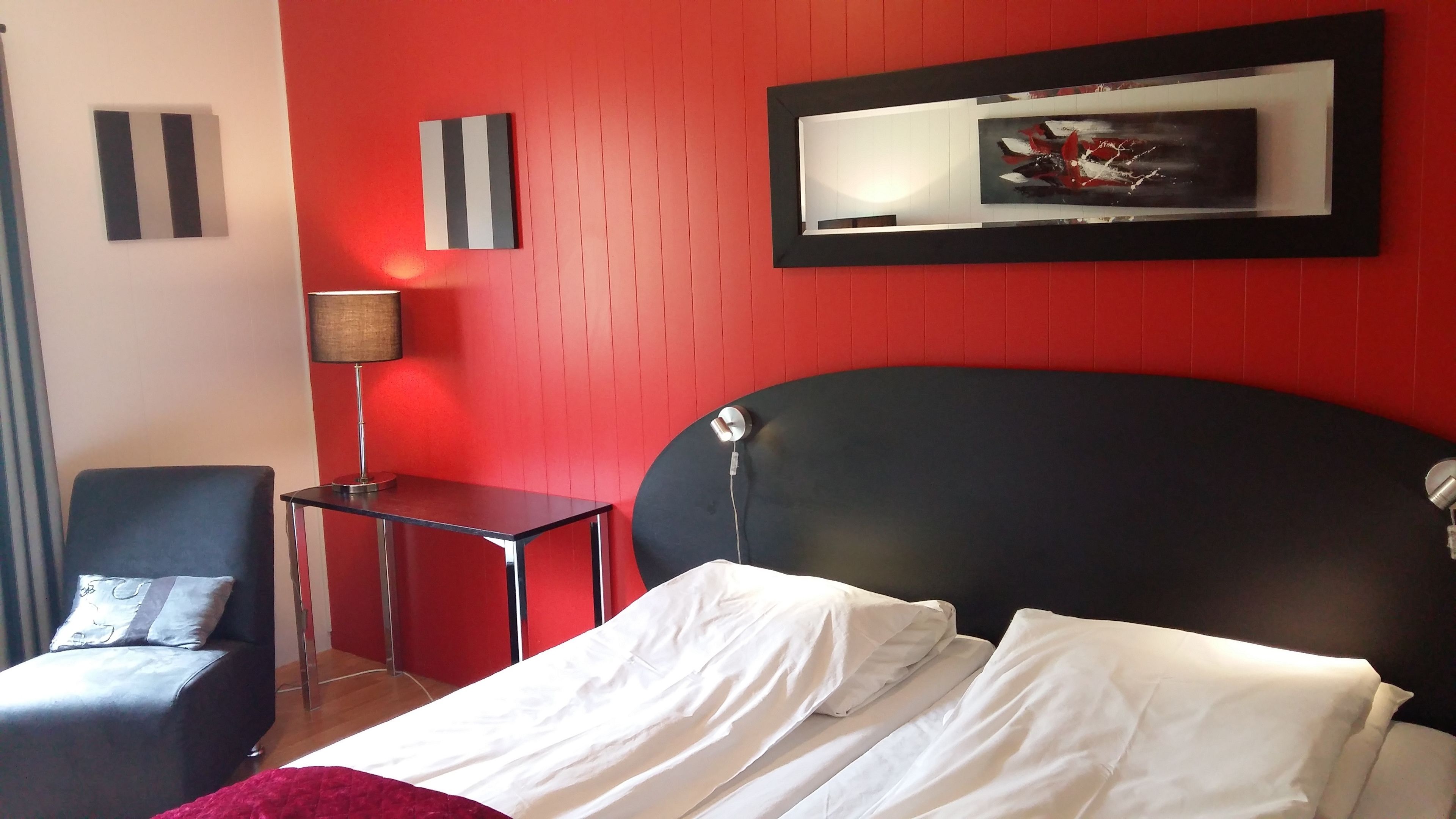 A hotel room with a bed, a desk, a chair, and a red wall.