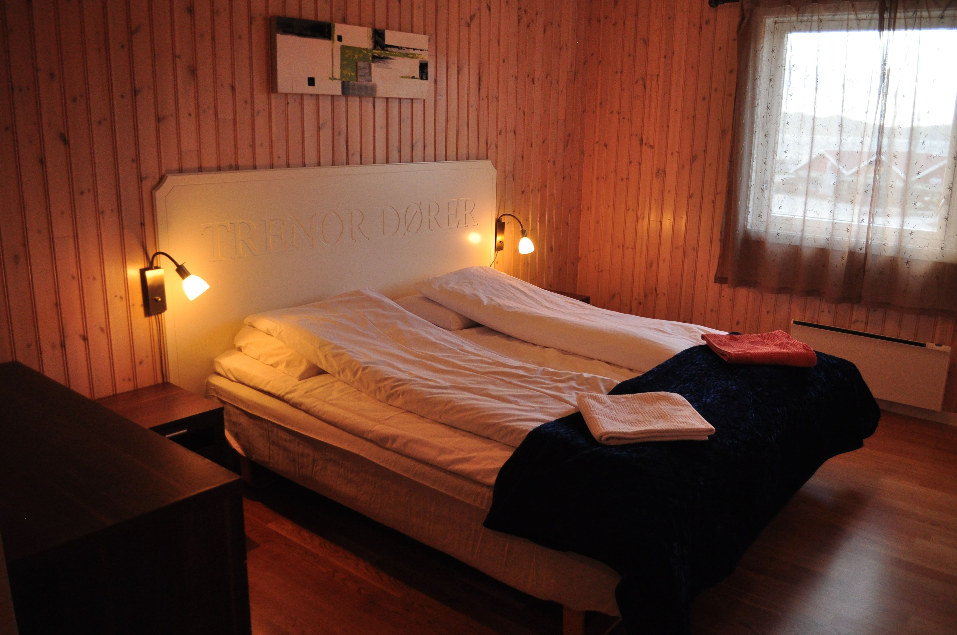 A bed with a headboard labeled "TRENOR DÖRER," two bedside lamps, a window with a sheer curtain, and a wooden floor.