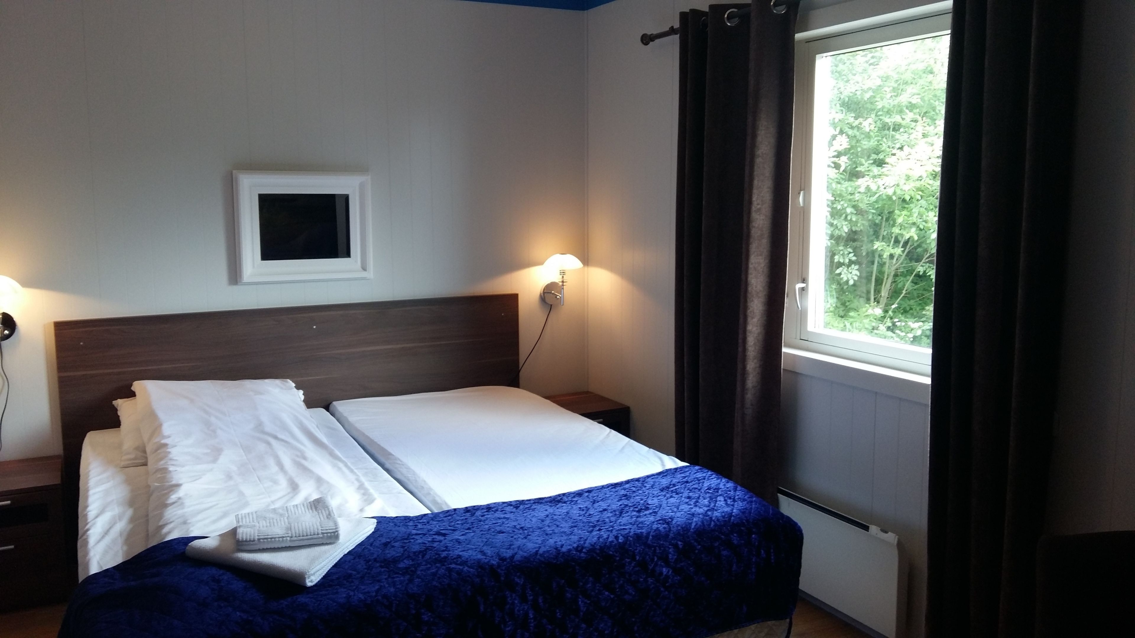 Un lit bien fait, avec du linge de lit blanc et une couverture bleue, une petite table de chevet avec une lampe, un tableau encadré au mur et une fenêtre avec des rideaux.