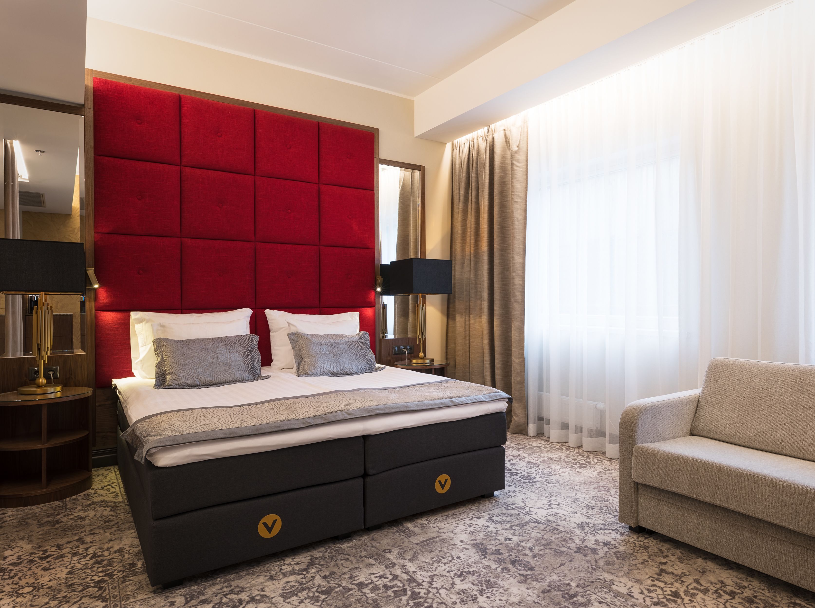 A hotel room with a large bed, a red headboard, a sofa, and a large window with curtains.