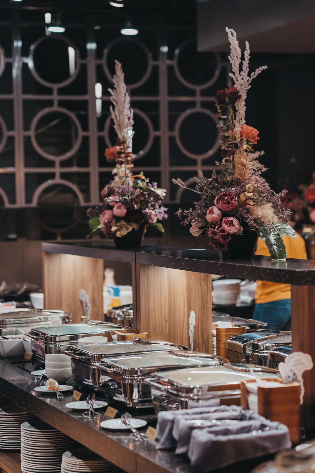 A buffet setup with multiple serving stations, a decorative floral arrangement, and a modern geometric wall design in the background.