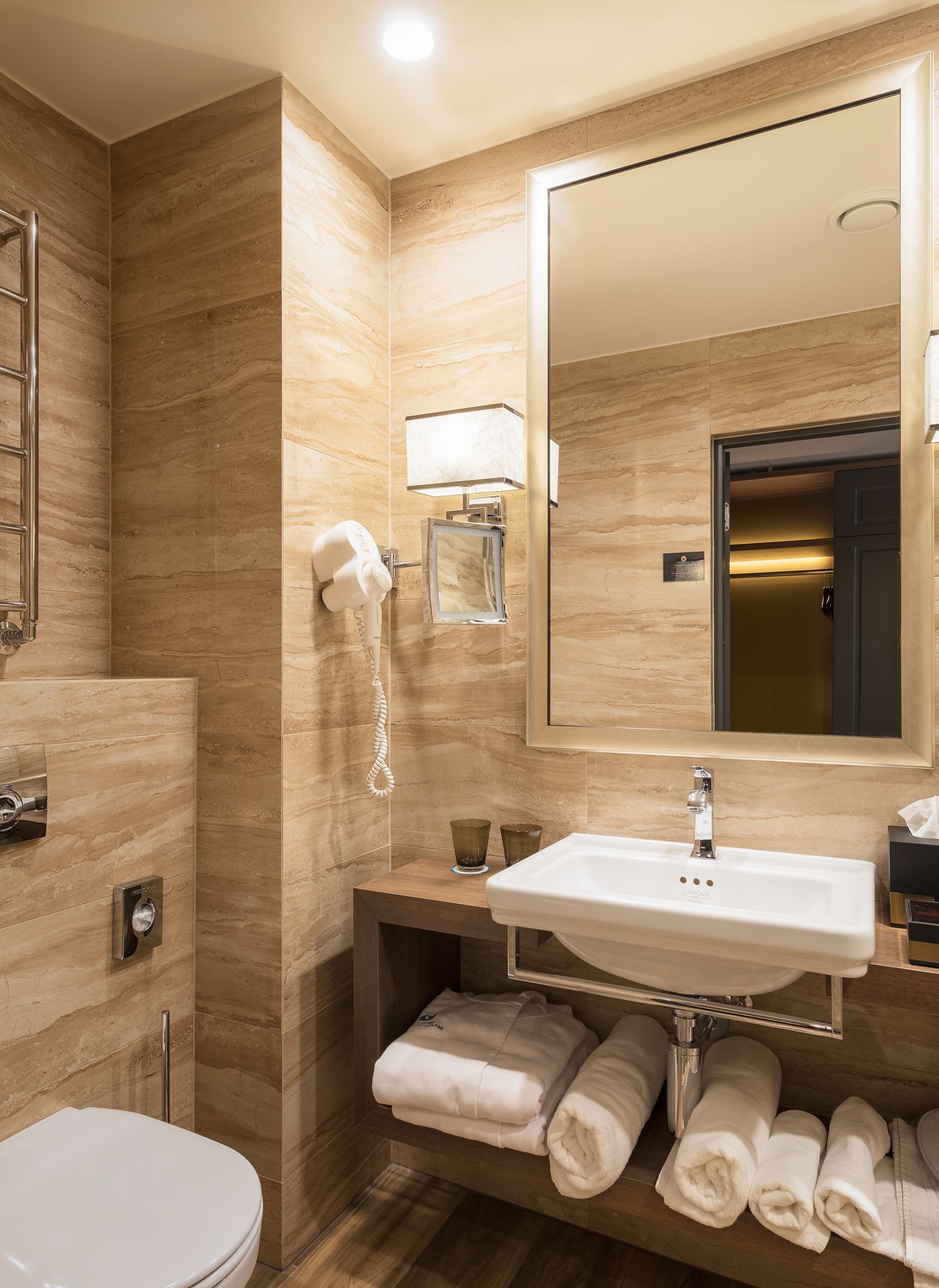 A modern bathroom with a marble wall, a large mirror, a sink, a hairdryer, and neatly folded towels.