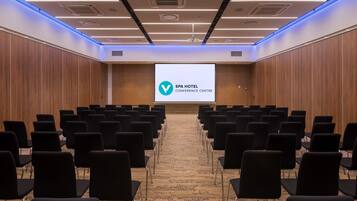 Un salón de conferencias con filas de sillas negras, una pantalla grande mostrando el logotipo del Centro de Conferencias Spa Hotel y paredes de paneles de madera.