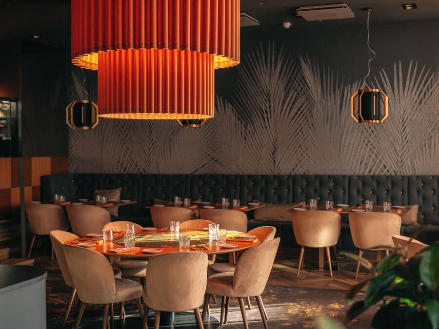 A modern restaurant interior with a central dining table surrounded by chairs, a unique red pendant light, and a patterned wall in the background.