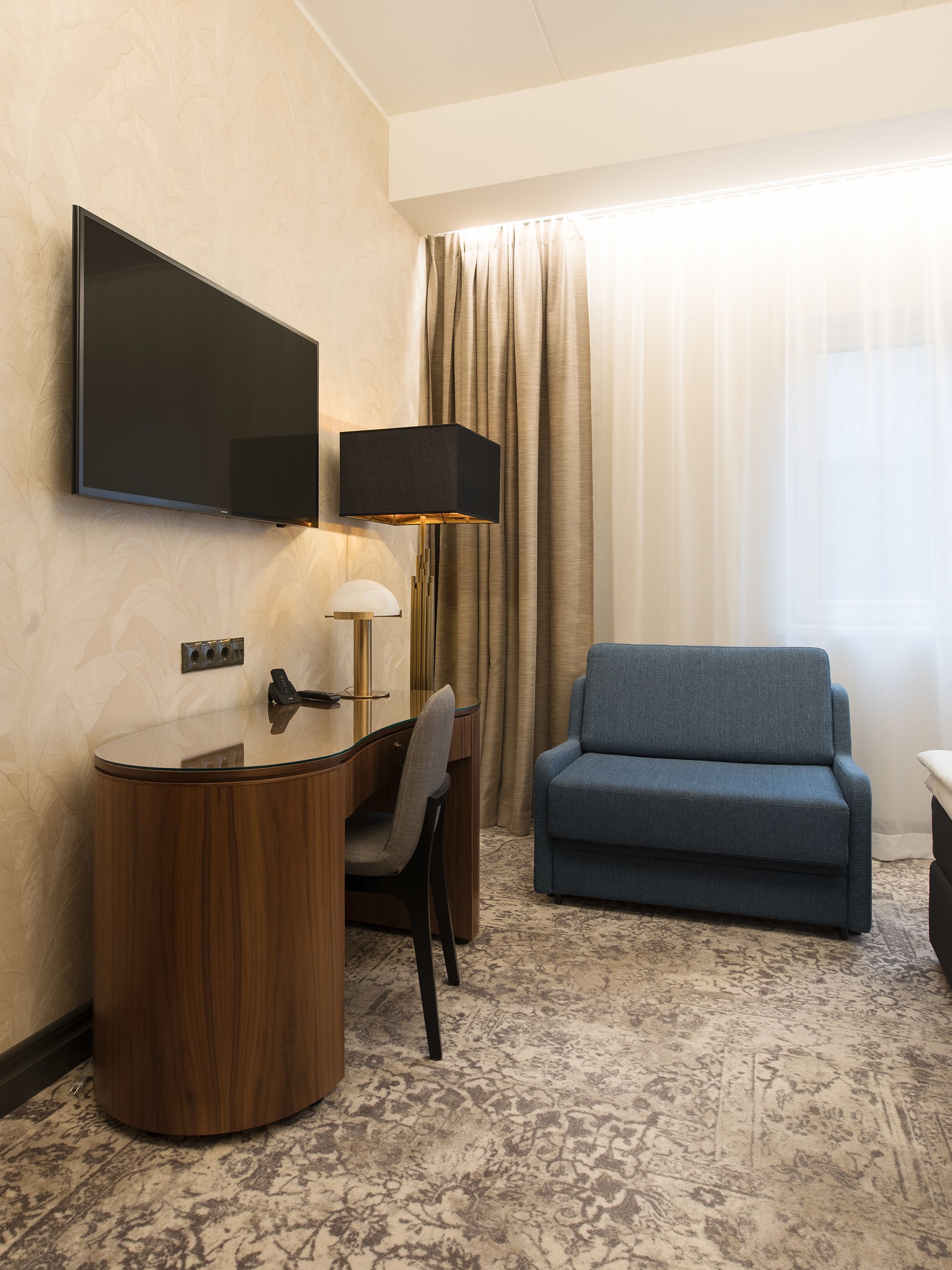 A hotel room with a desk, chair, sofa, and a flat-screen TV mounted on the wall.