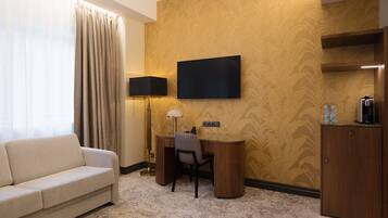 Quarto de hotel com sofá, escrivaninha, televisão de tela plana, luminária e minibarra.