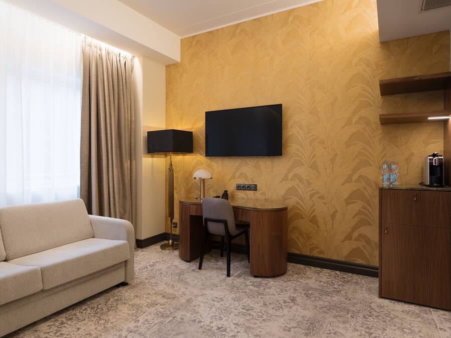 Quarto de hotel com sofá, escrivaninha, televisão de tela plana, luminária e minibarra.
