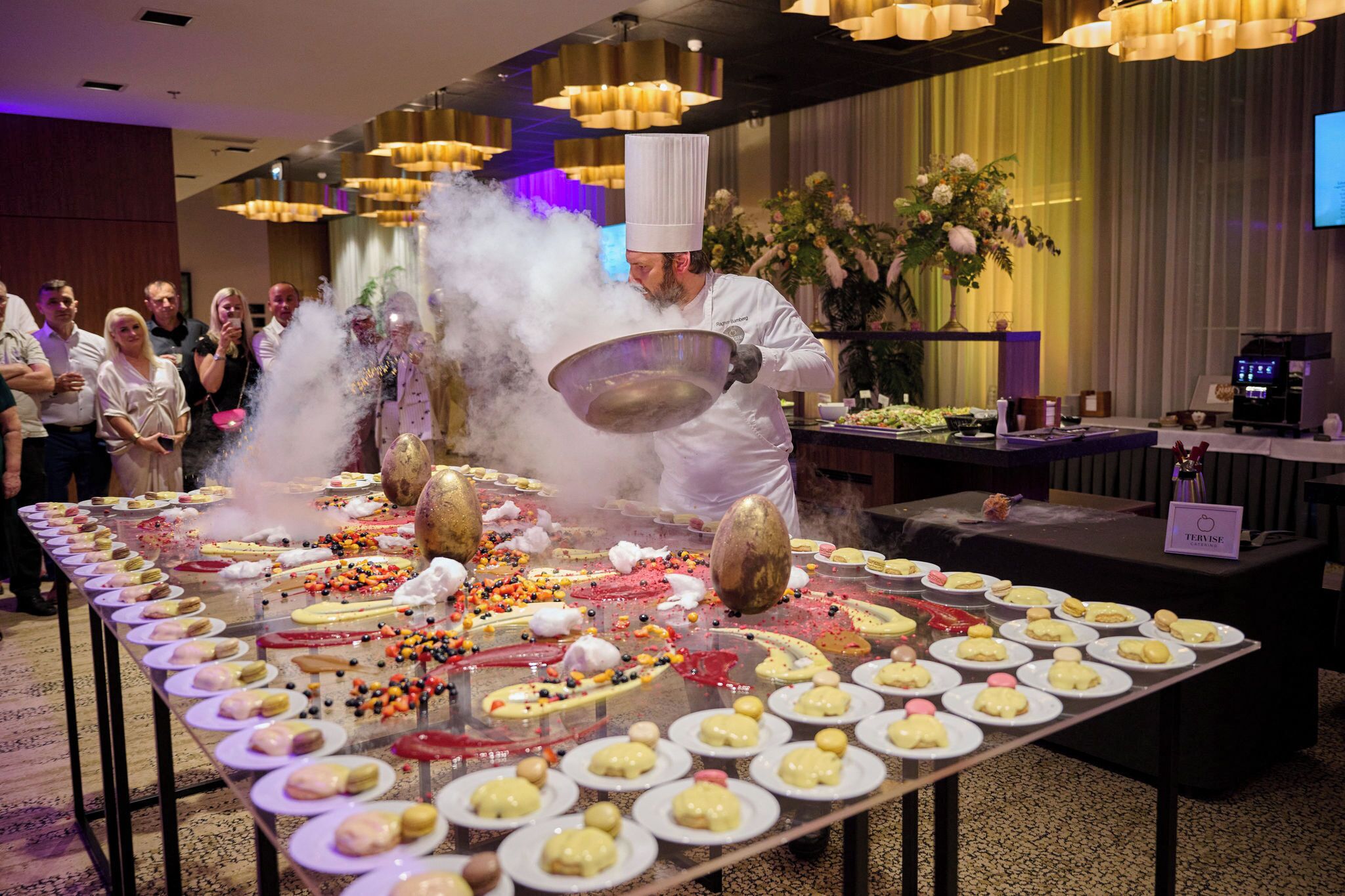 Un chef présente une démonstration culinaire à l’aide d’un grand wok dans une pièce bien éclairée, devant une longue table remplie de plats.