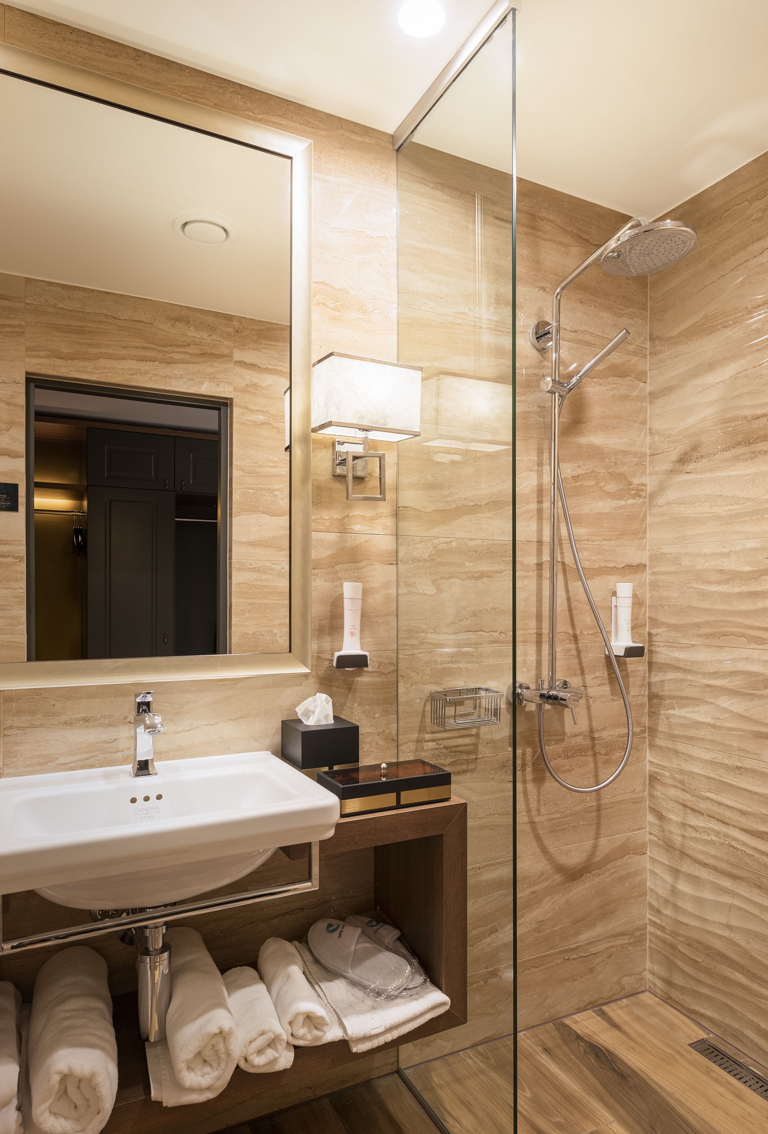 Uma casa de banho moderna com chuveiro em cabine de vidro, um espelho grande, uma pia com porta-toalhas em baixo e um pequeno armário com produtos de higiene pessoal.
