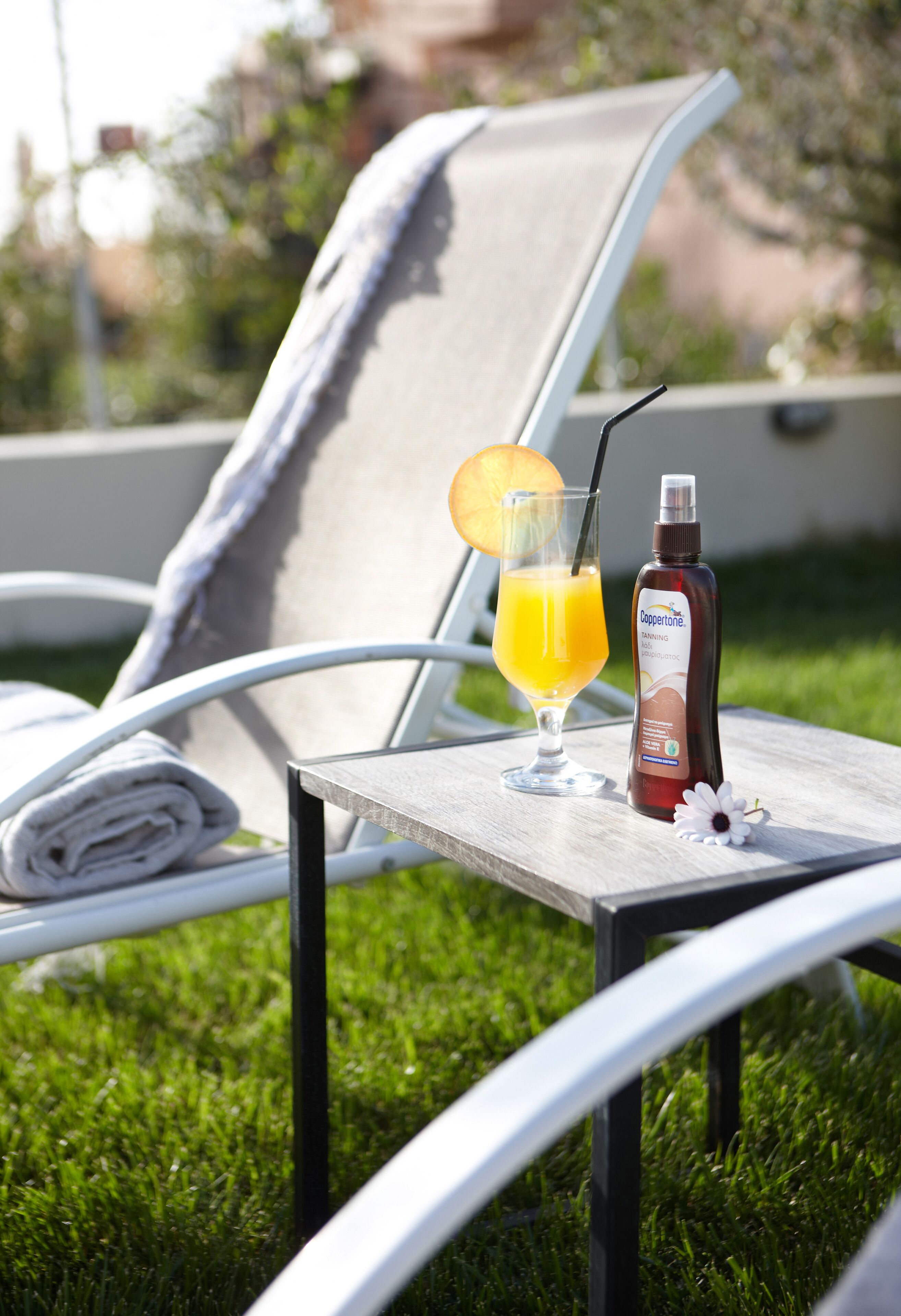 Une chaise longue, une serviette, un verre de jus d’orange avec une paille et une bouteille de Cuppette sur une table en bois.