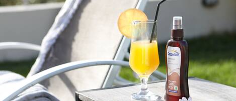 Une chaise longue, une serviette, un verre de jus d’orange avec une paille et une bouteille de Cuppette sur une table en bois.