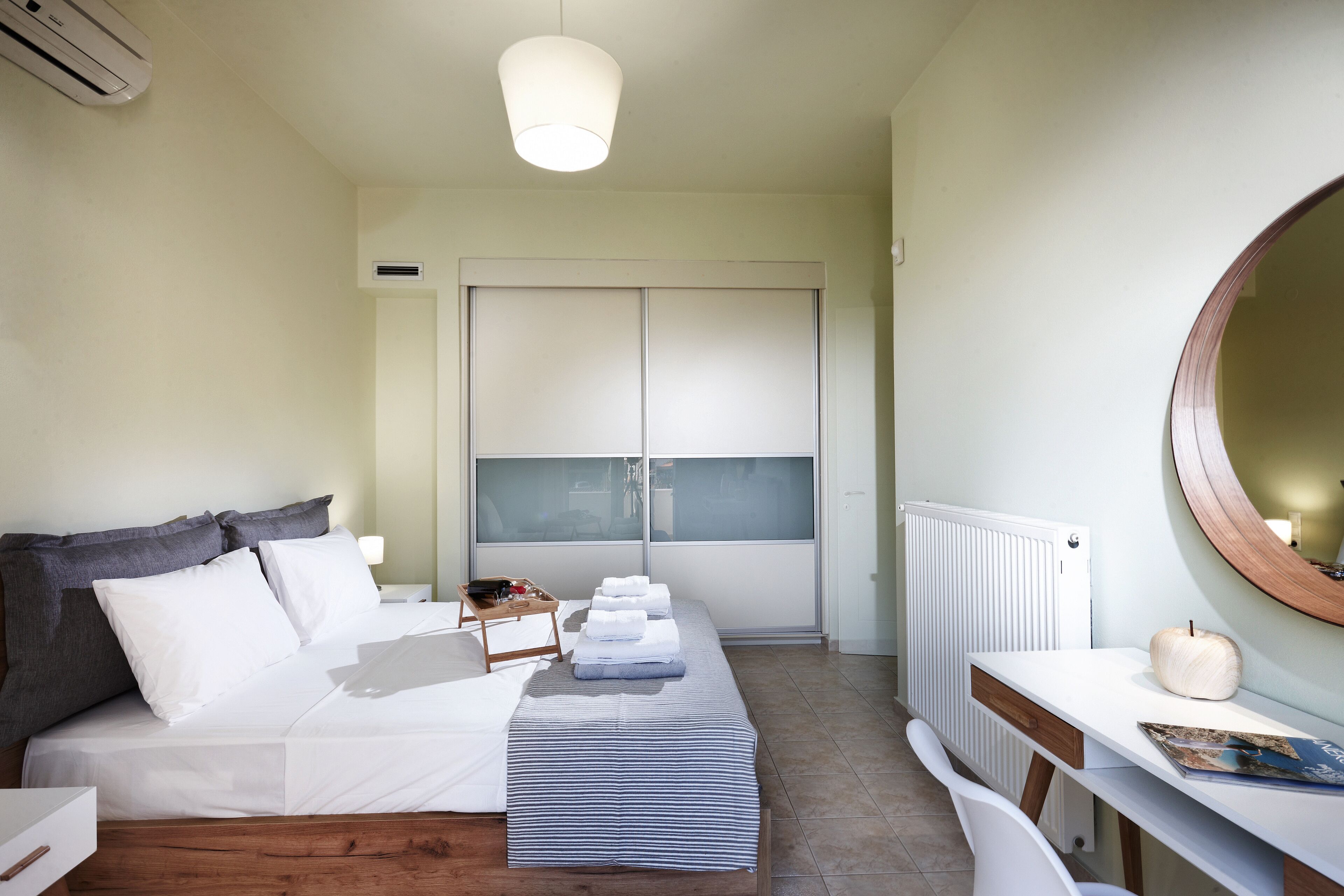 Ein ordentlich bezogenes Bett mit weißen Leinen, ein Nachttisch mit Lampe, ein Holztisch mit Spiegel und ein Fenster mit Jalousien.