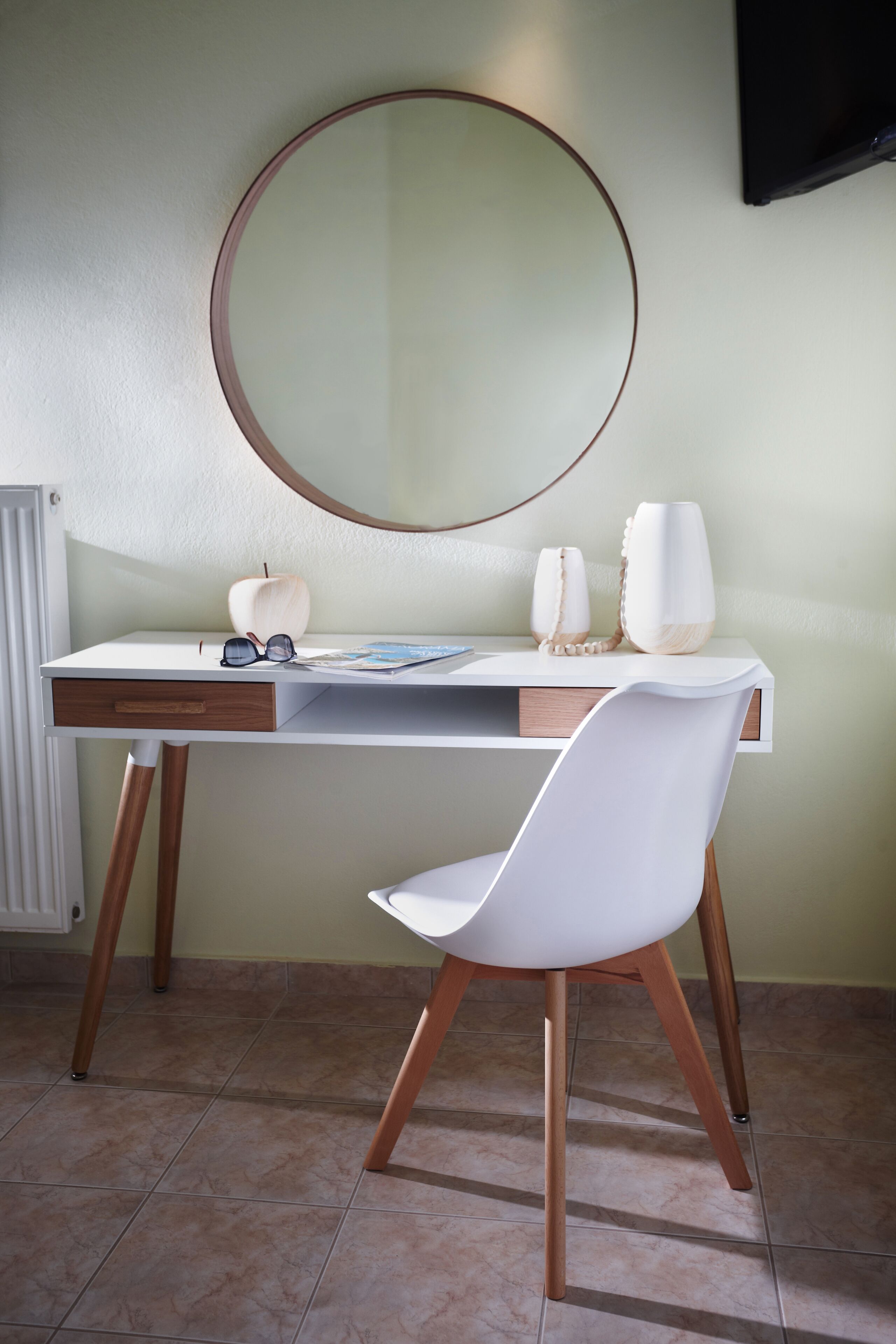 A modern desk with a white chair, a round mirror, and two white vases on the desk.