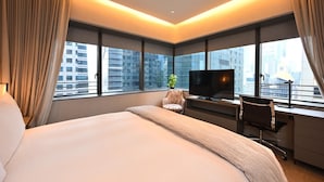 Habitación de hotel moderna con una cama grande, un escritorio con computadora, televisión y vistas de la ciudad a través de amplios ventanales.