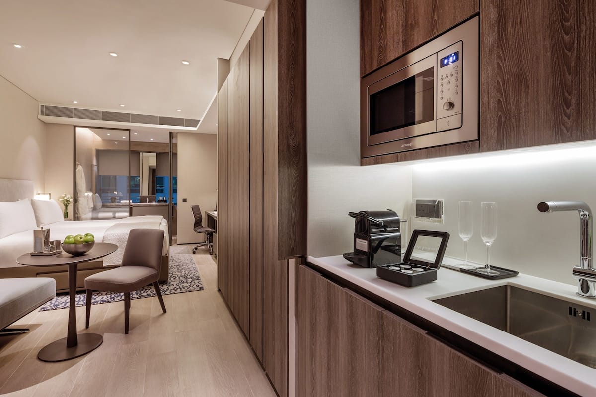 A modern hotel room with a kitchenette, a bed, a seating area, and a view of the city.