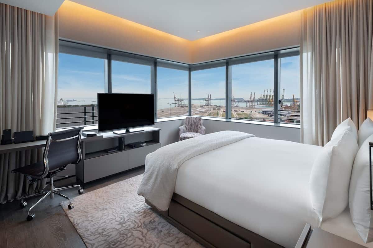 A hotel room with a large bed, a desk with a flat-screen TV, and a view of a port with cranes.