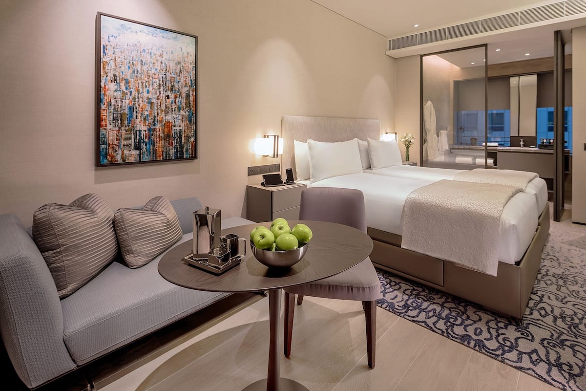 A modern hotel room with a bed, a sofa, a table with a bowl of apples, and a large abstract painting on the wall.