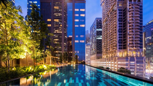 Un hotel moderno urbano con una piscina reflectante, edificios altos y abundante vegetación.
