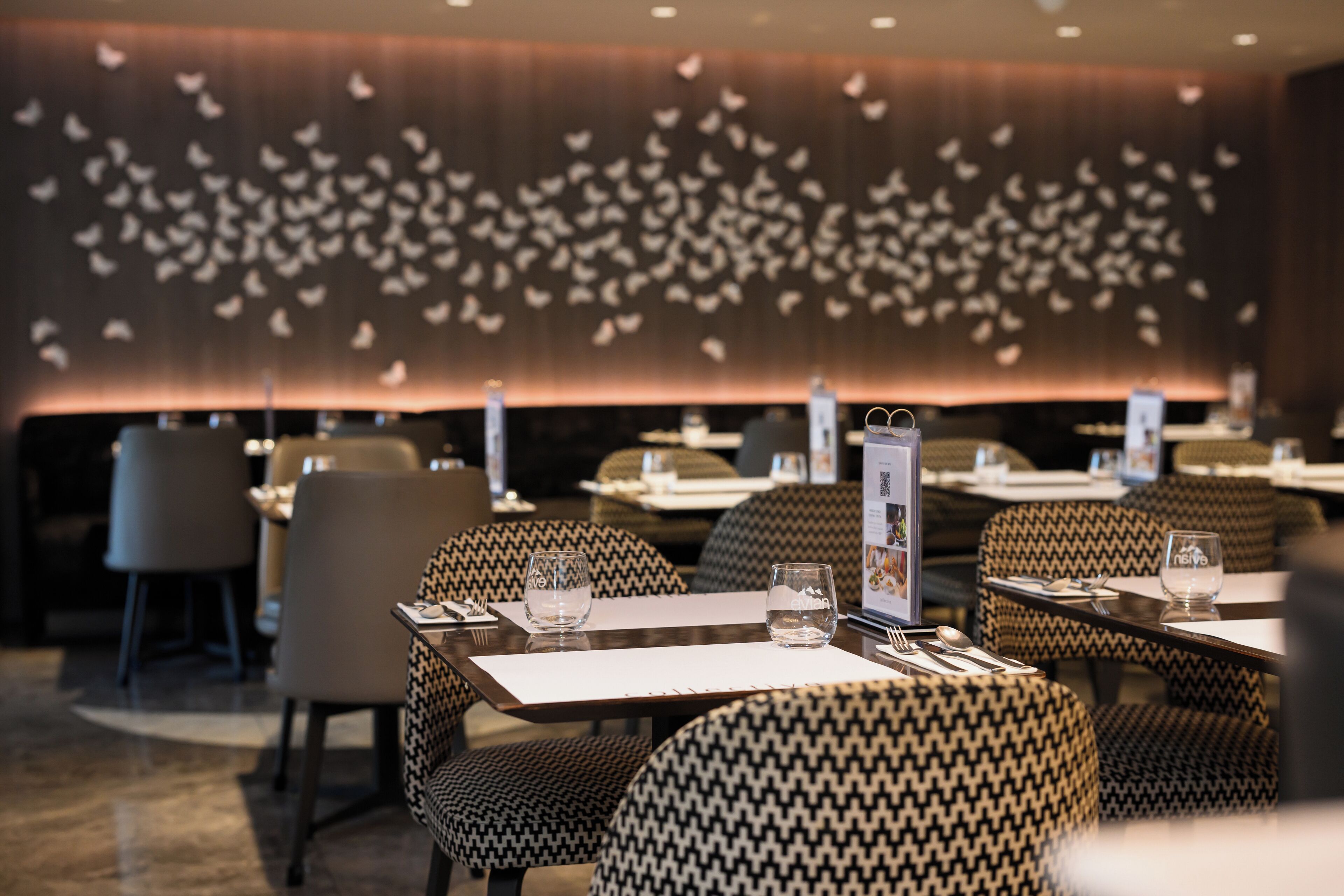 L’intérieur d’un restaurant présente des chaises à motifs pied-de-poule, des tables avec des menus et un mur décoré d’un groupe d’oiseaux blancs.