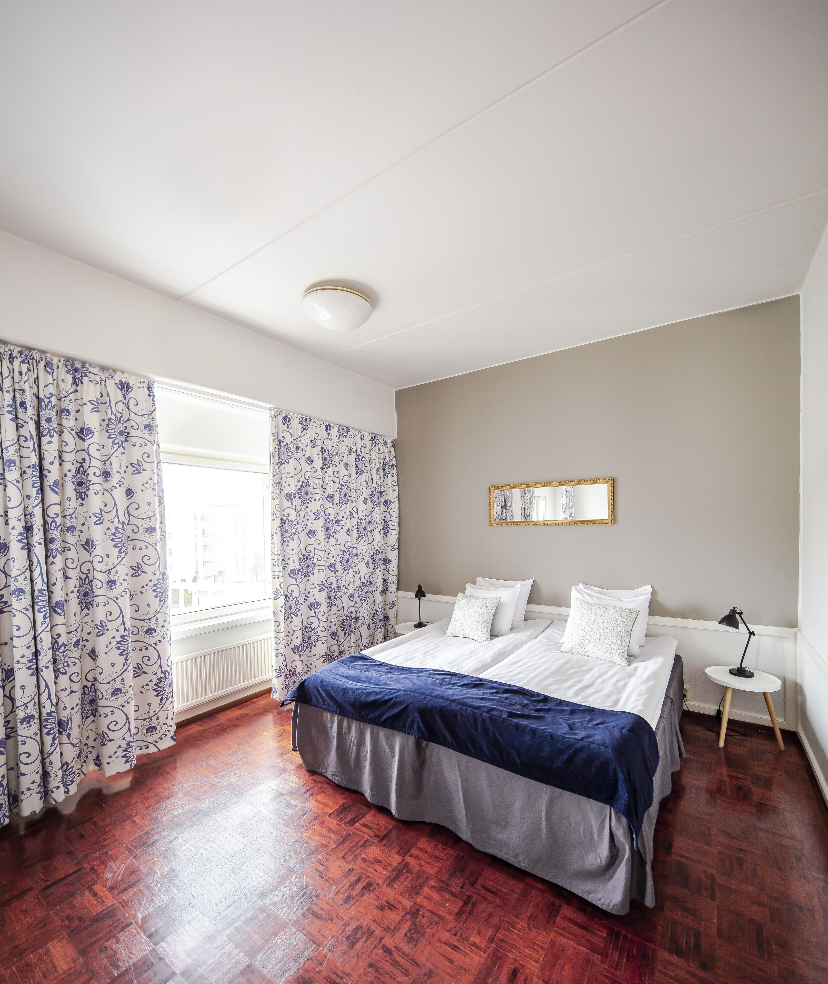 Un dormitorio con una cama doble, una mesita de noche con lámpara, una ventana con cortinas y un radiador.