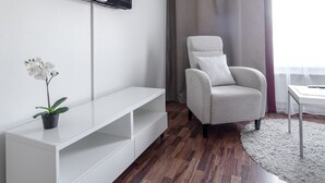 Un salon moderne avec un téléviseur à écran plat fixé au mur, un meuble de rangement blanc, un fauteuil gris et une petite table avec une télécommande.