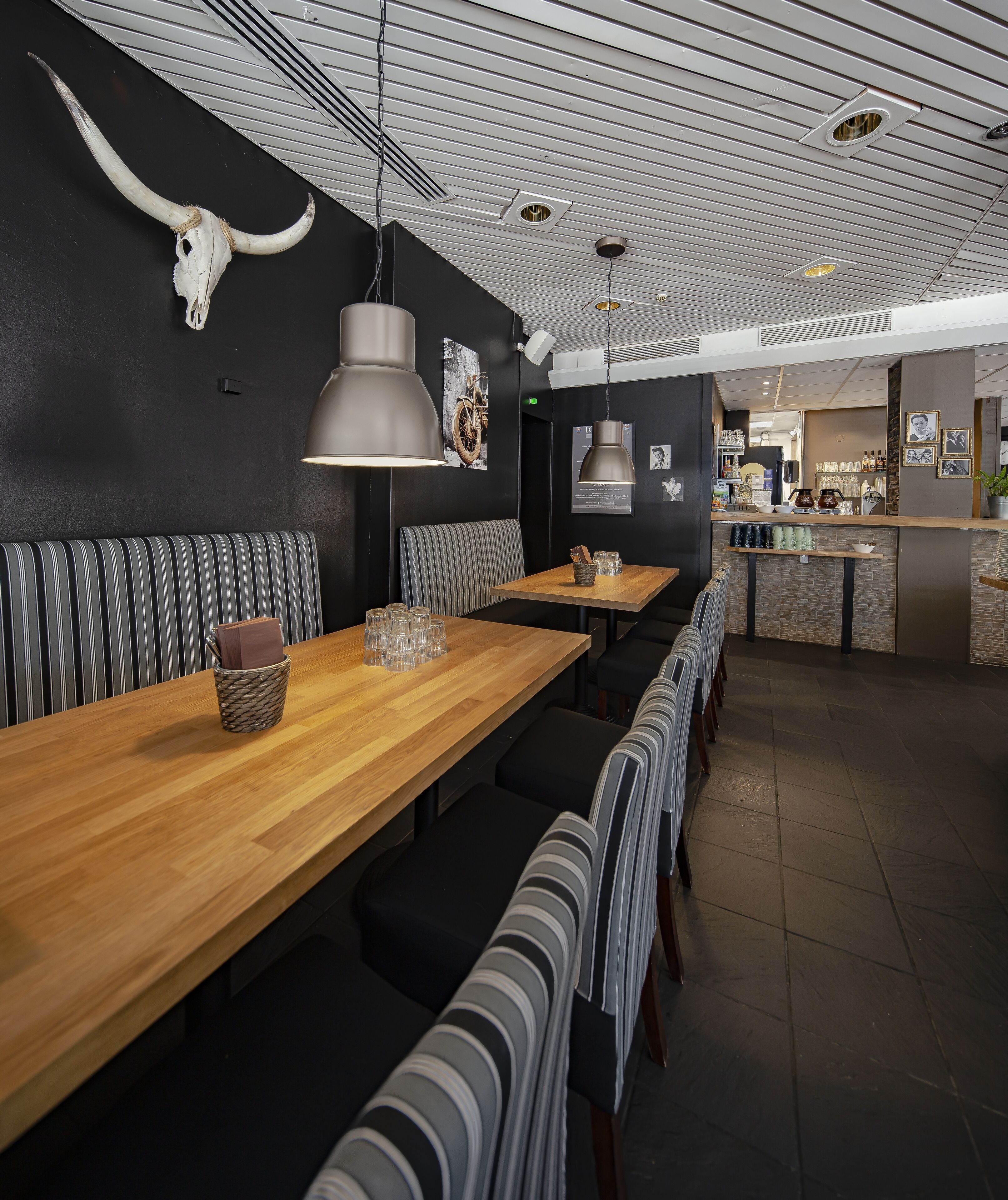 A restaurant interior with long wooden tables, striped booths, and hanging pendant lights.
