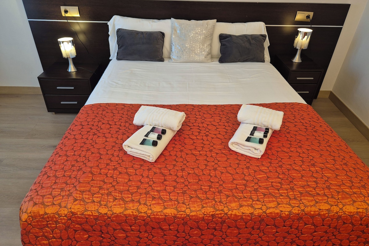 Una cama bien hecha con una colcha estampada roja, dos toallas dobladas y dos lámparas de noche.
