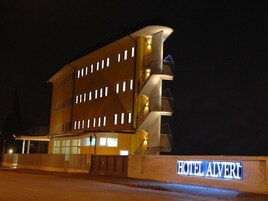 A multi-story hotel with a prominent balcony on the right side, a well-lit entrance, and a sign reading "HOTEL ALVERI".