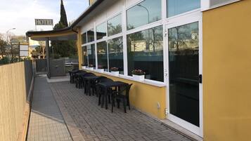 Exterior de un hotel con fachada amarilla, ventanas grandes, área de estar al aire libre con mesas y sillas, y un cartel que indica el nombre del hotel.