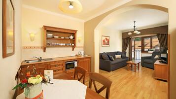 Un comedor con mesa de madera, sillas y un centro de mesa decorativo. Se aprecia una zona de cocina con armarios de madera y un fregadero. En el fondo, se observa una sala de estar con sofá y una mesita.