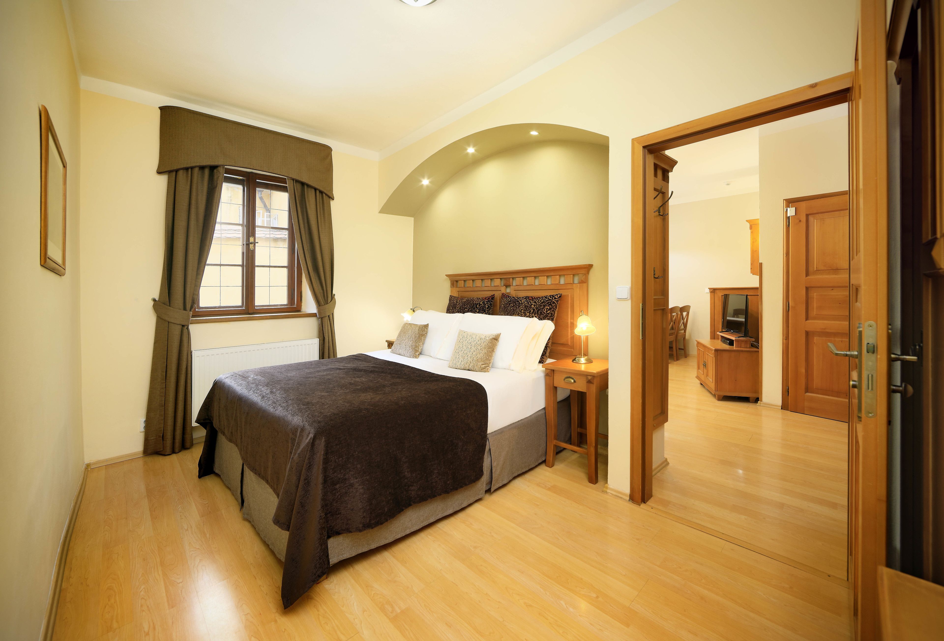 A bedroom with a bed, a nightstand, a window with curtains, and a wooden door leading to another room.